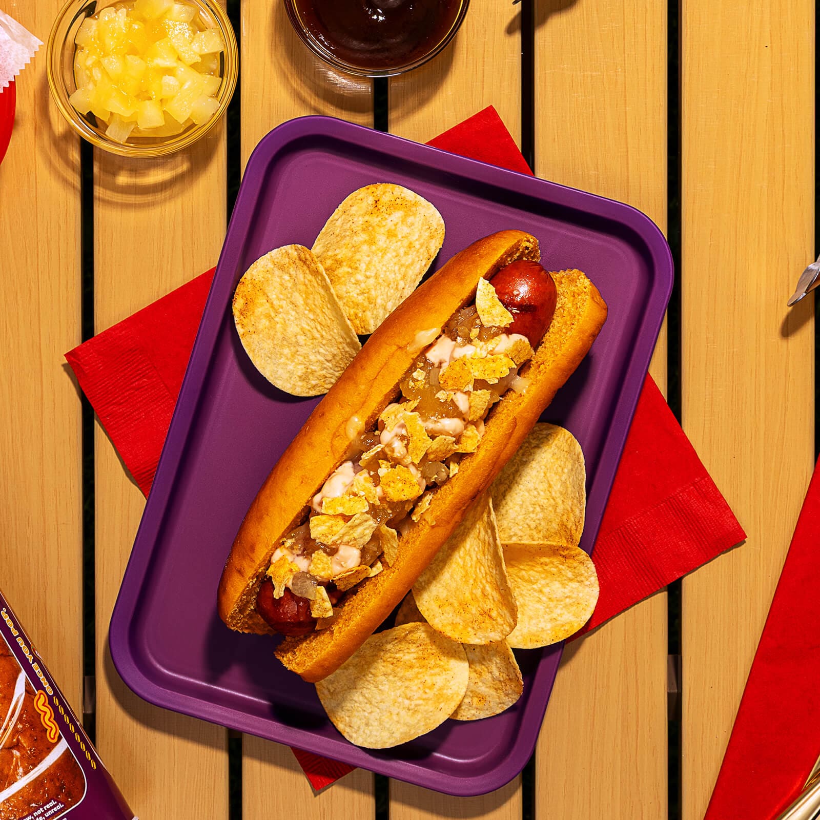 A cooked hot dog in a Pringles BBQ bun, topped with melted mozzarella, pineapple jam, pink sauce, and crushed Pringles BBQ potato crisps, served on a purple tray.